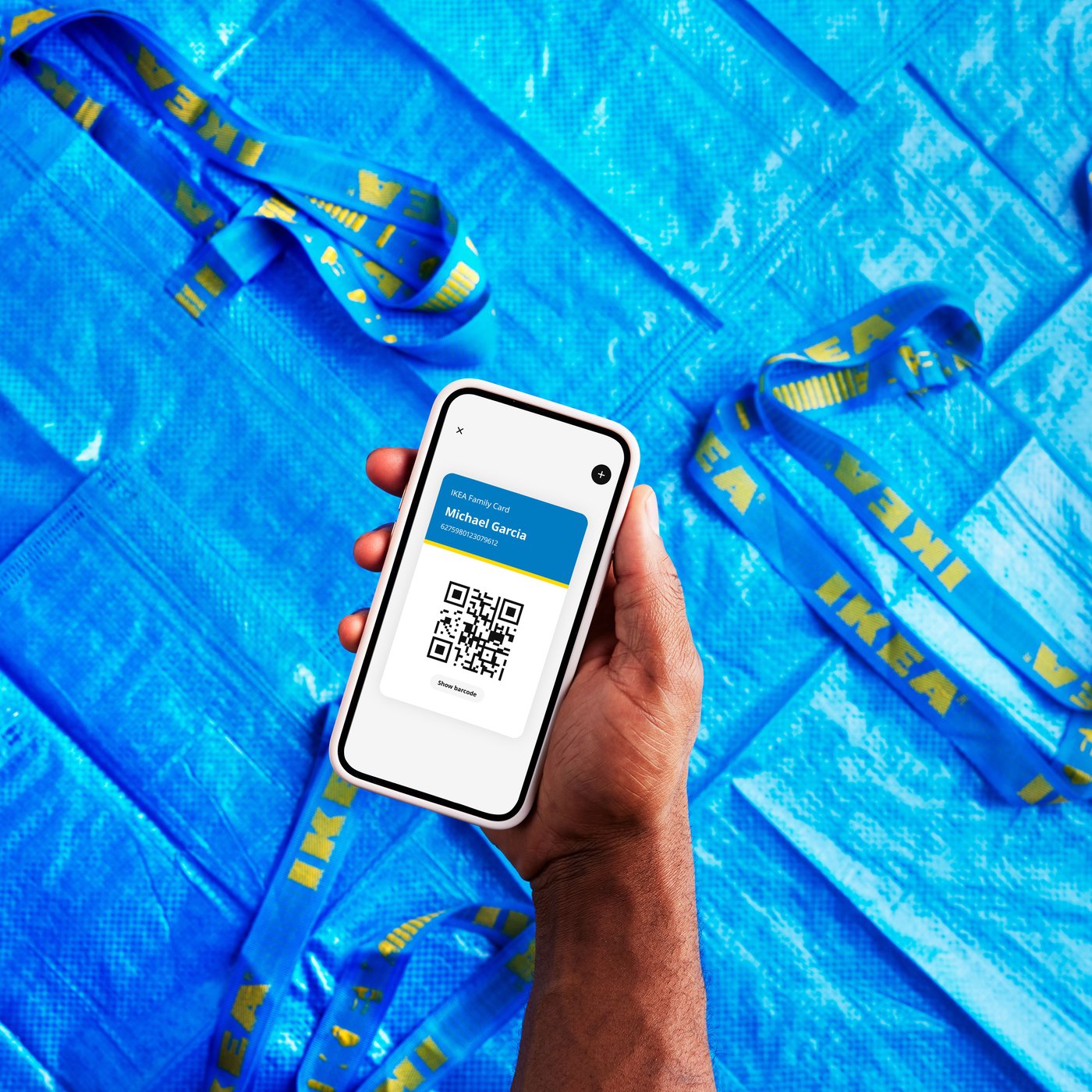 A hand holding a smartphone displaying the IKEA app above a blue background with several blue FRAKTA bags.