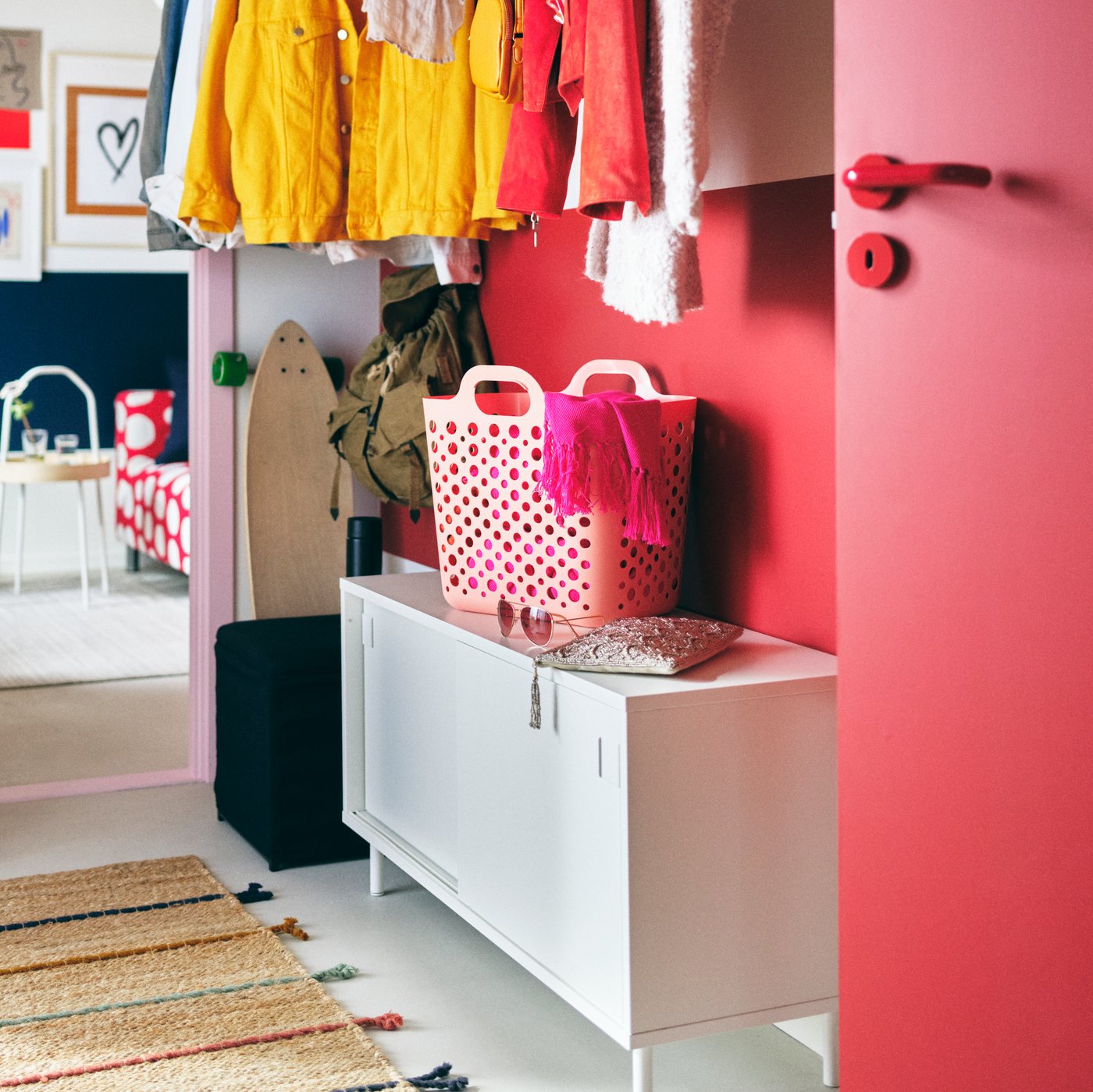 Una entrada de un hogar, con paredes color rosa. Un mueble  blanco TJILLEVIPS, que tiene un canasto color rosa claro SLIBB. En el piso una alfombra RAKLEV. 