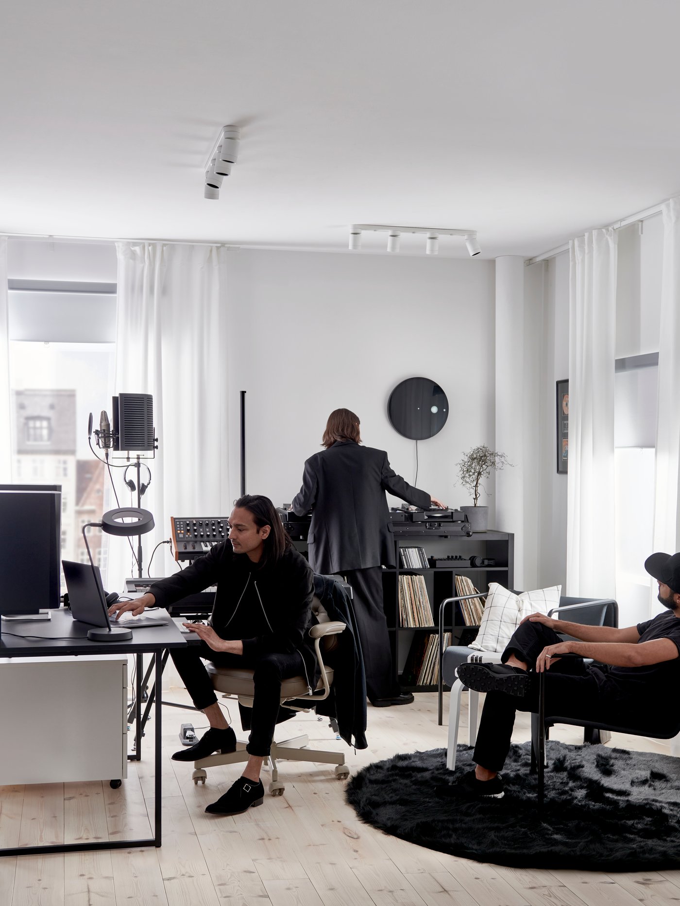 Three people dressed in black in a white room with black OBEGRÄNSAD rug, desk, shelving unit and armchair.
