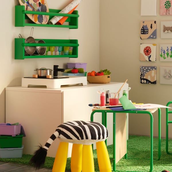 A STENMUS mini play kitchen on a SMÅSTAD bench with toy storage with FLISAT wall storage above it in the corner of a room.