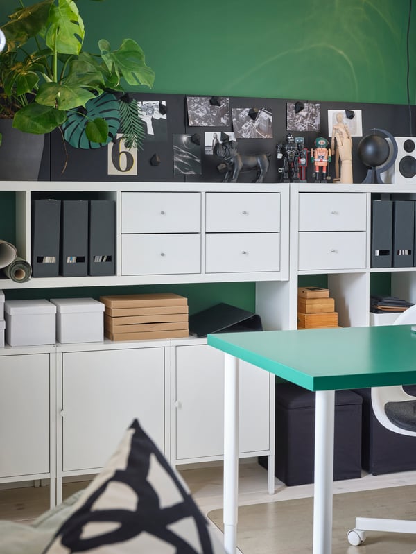 A green wall with white shelving units, drawers and cabinets in front. Boxes and files are on the shelves.