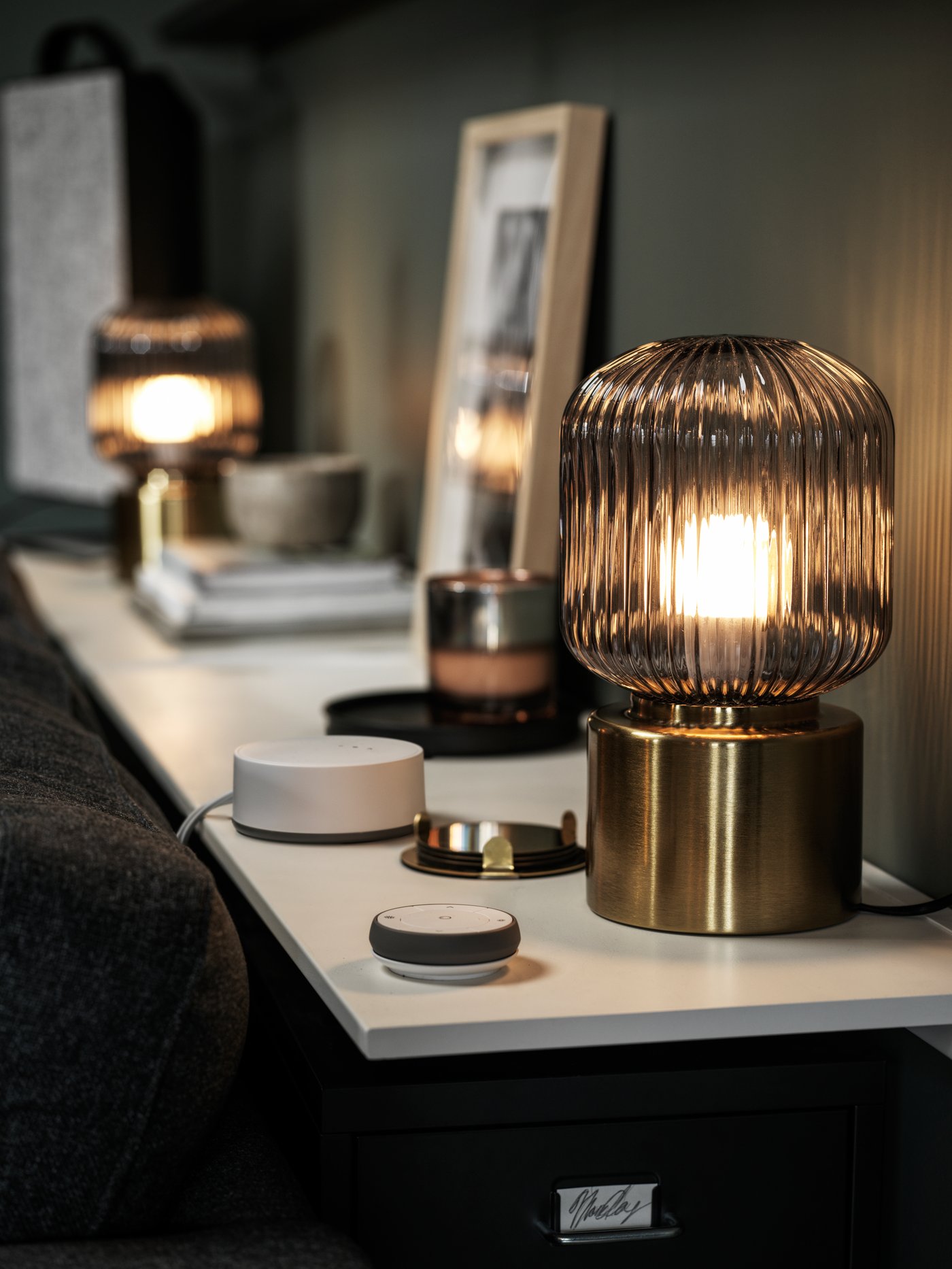 A wall shelf behind a sofa holds SOLKLINT brass table lamps, a LIVBOJ charger, a TRÅDFRI remote control and decoration.