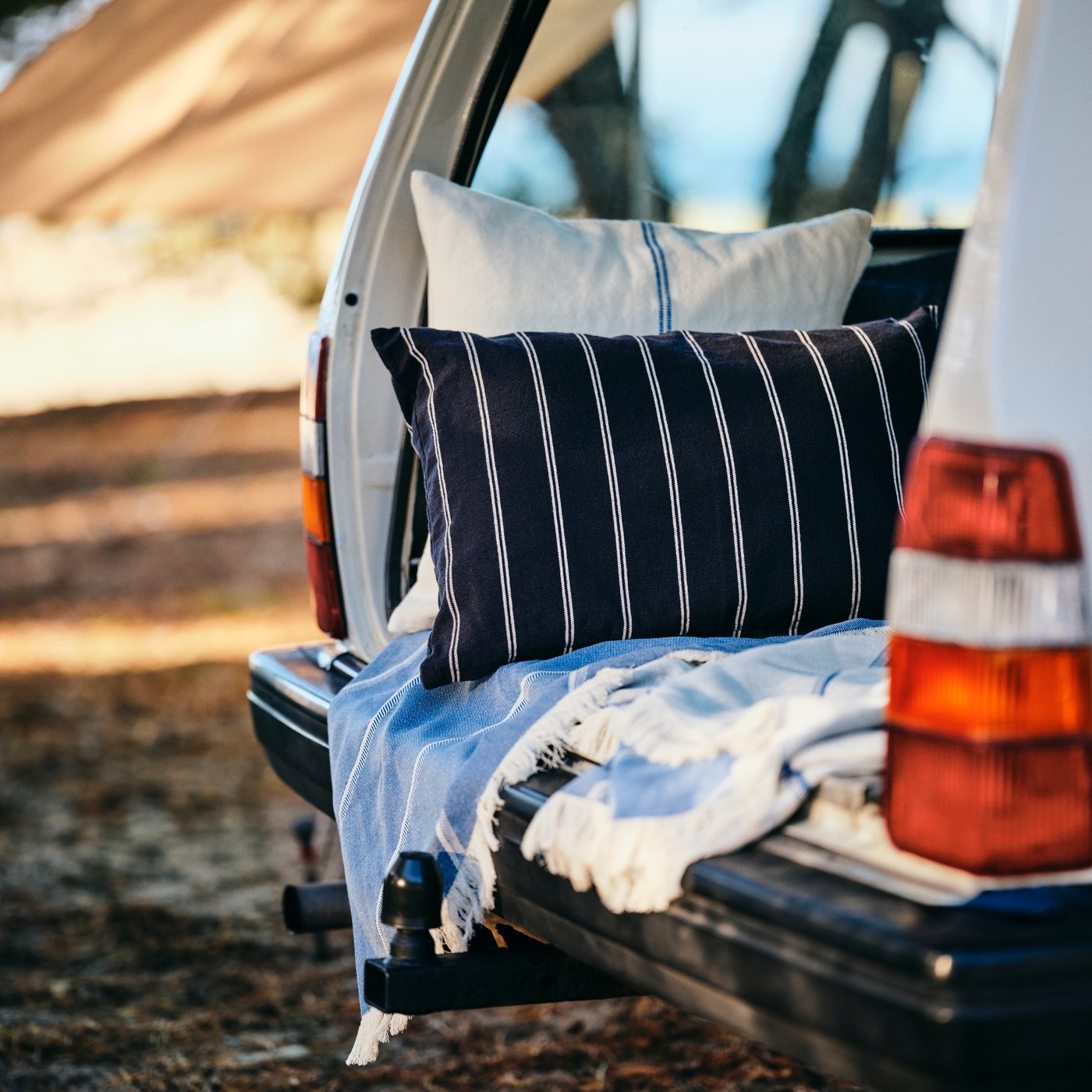 Two pillows with SOLUPPGÅNG cushion covers and a SOLUPPGÅNG throw form a cosy nook in the back of a car.