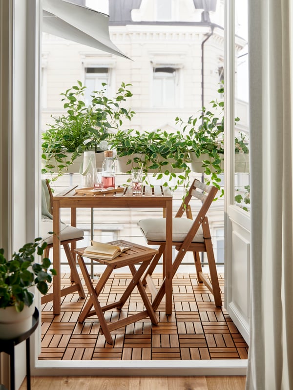 A light-brown-stained NÄMMARÖ outdoor table, a foldable stool and foldable chairs on a balcony enveloped in leafy plants.