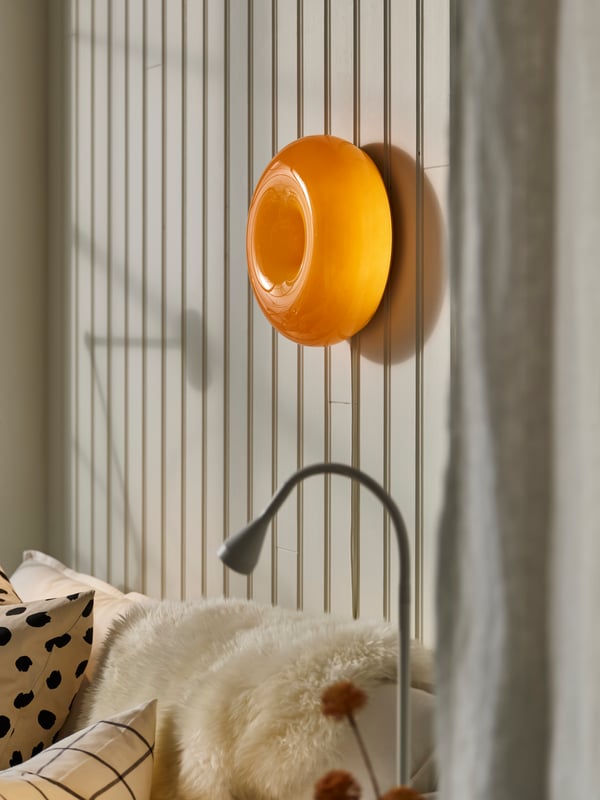 Lampu dinding VARMBLIXT bulat berwarna jingga di sebelah atas sofa berwarna putih dengan bulu kambing ULLERSLEV berwarna putih pudar, serta lampu kerja NÄVLINGE berwarna putih.