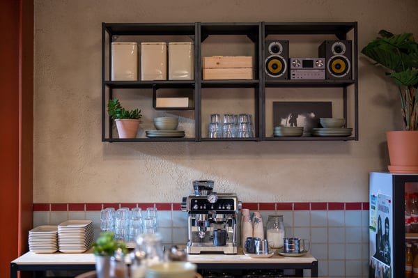 Een IKEA keukenwand met opbergmodules en open planken, gecombineerd met servies, een koffiemachine en planten, vormt een praktische en gezellige koffieruimte.