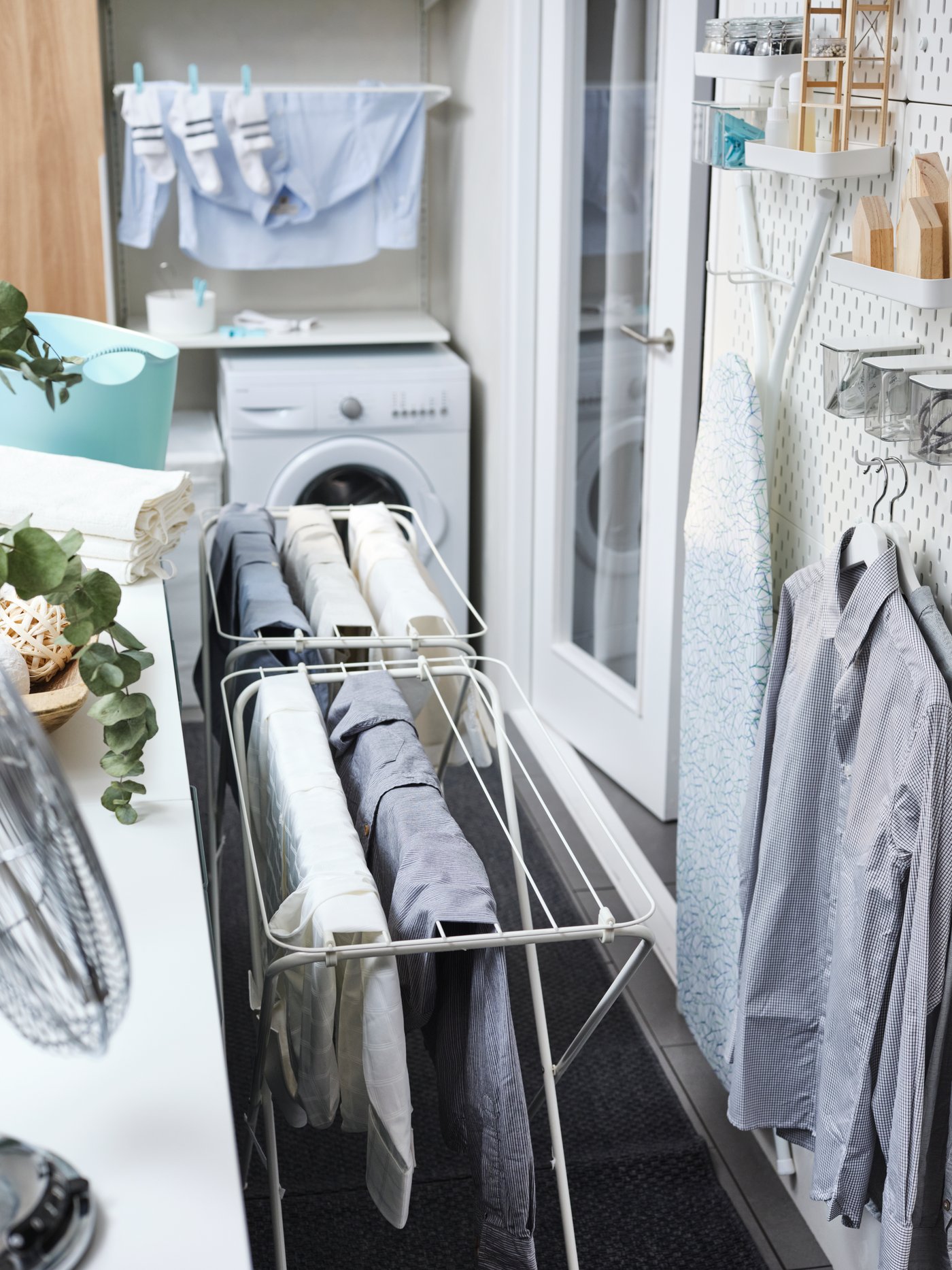 Es sind zwei weiße JÄLL Wäschetrockner mit Kleidung zu sehen, die neben einem weißen Regal und Wandaufbewahrung mit Hemden an Bügeln und einem Bügelbrett stehen.