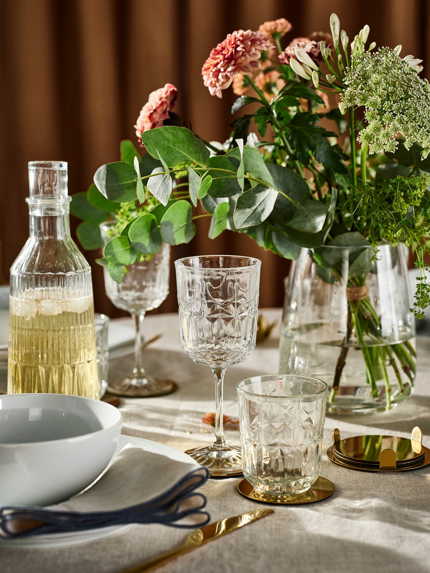 En SÄLLSKAPLIG glaskaraffel, glas og vinglas, plus blomster i en glasvase, på et bord med en naturfarvet/beige dug.