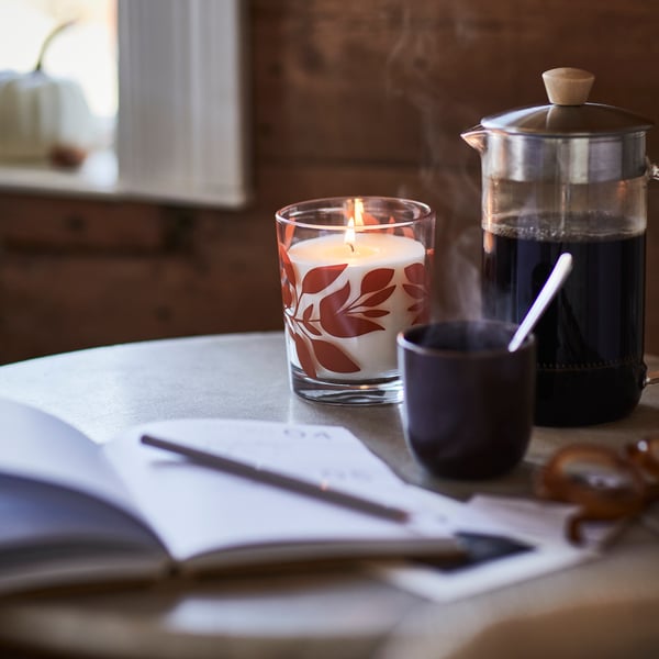 A HÖSTAGILLE scented candle in a glass sits on a table beside a French press and a steaming mug.
