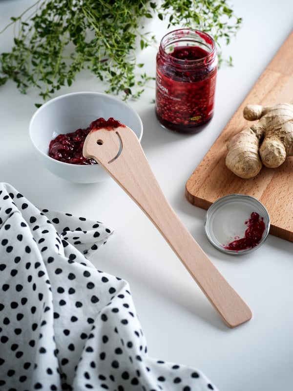 Beech UPPLIVAD spoon shaped like a smiley face balances on the side of a bowl with red SYLT LINGON spread on a counter.