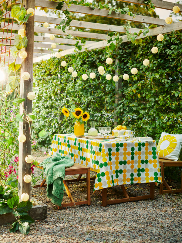 An outdoor table made with a dot-patterned BRÖGGAN tablecloth and tableware, surrounded by greenery and a lighting chain.
