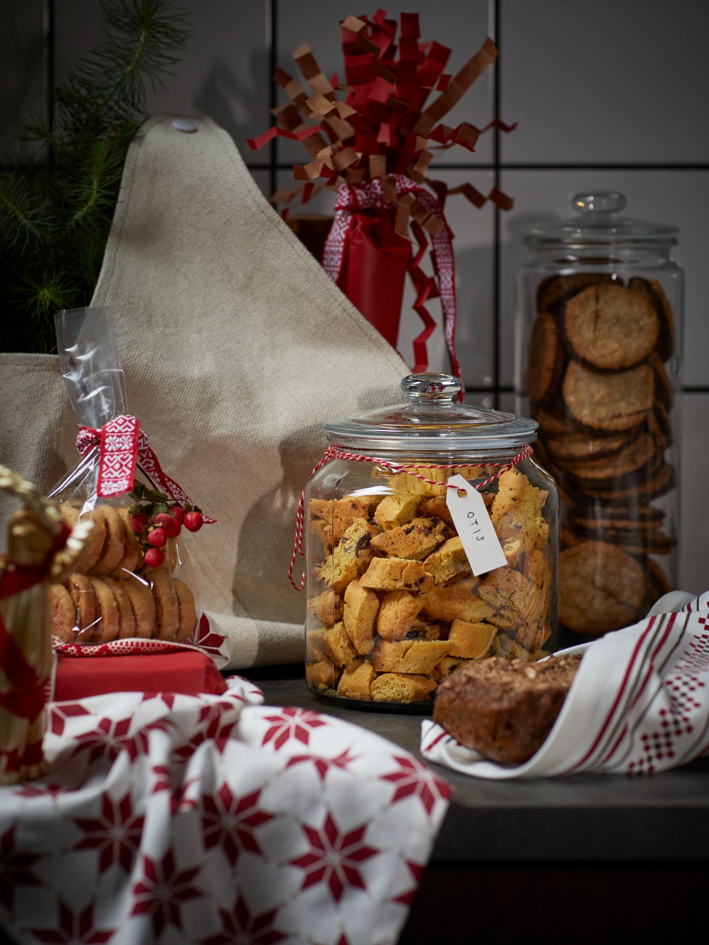 A cookie-filled VARDAGEN glass jar, surrounded by a bread loaf wrapped in a VINTER 2021 tea towel and other baked goodies.