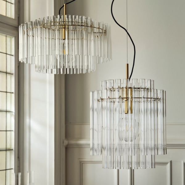 Two brass-plated STOCKHOLM 2025 pendant lamps with glass tubes hang side by side by a large grid window.