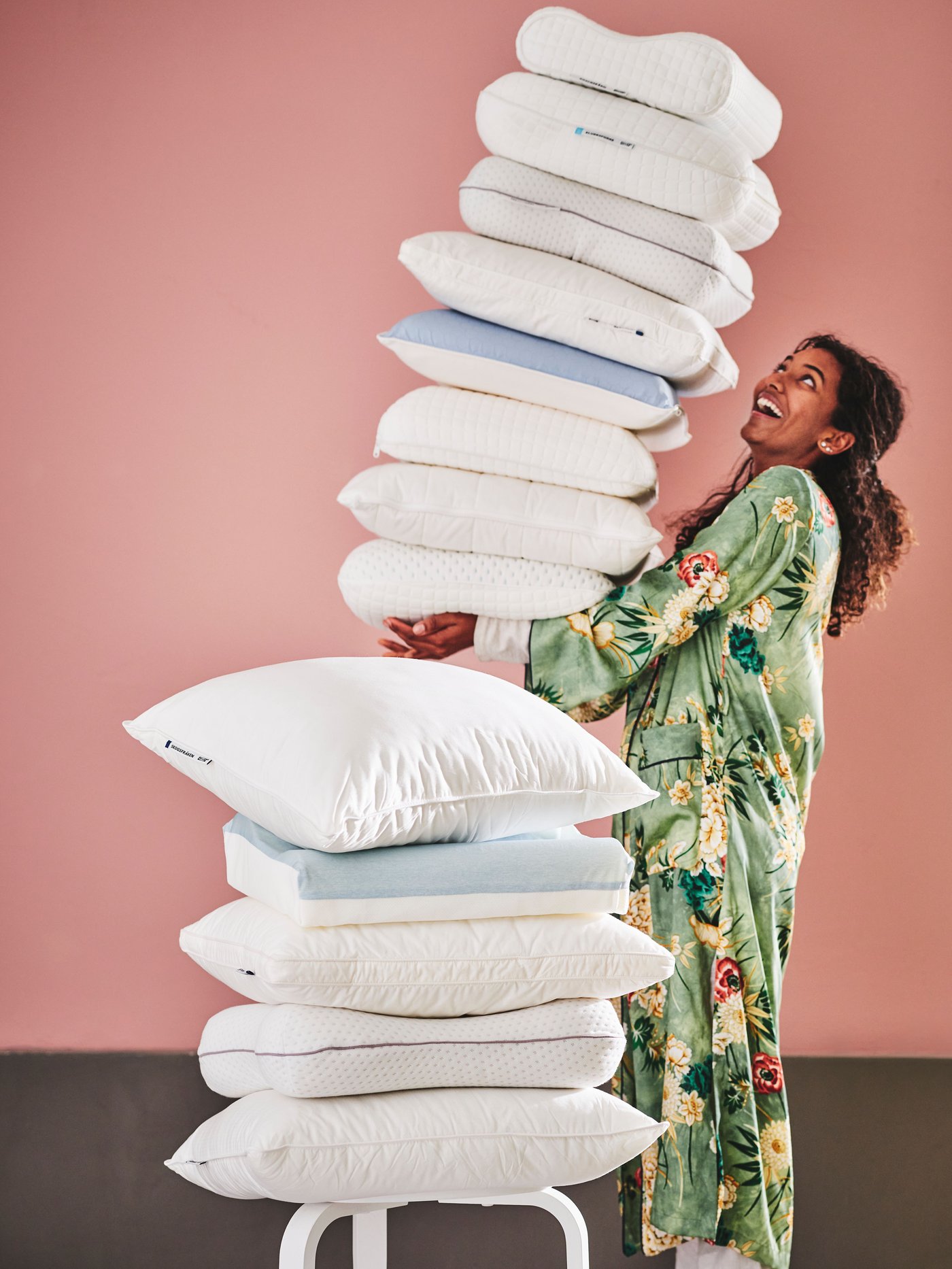 Una mujer cargando una torre de almohadas IKEA y al costad de ella una silla con más almohadas.
