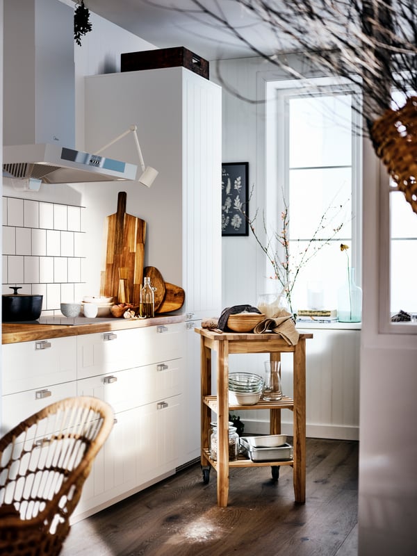 A country-style kitchen with a SÄLJAN wooden worktop and white cabinets, and a birch BEKVÄM trolley filled with cookware.