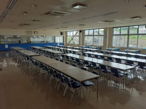 長テーブルと青い椅子が規則的に並ぶ広い食堂スペース。大きな窓から光が入り、奥に配膳カウンターが見える。