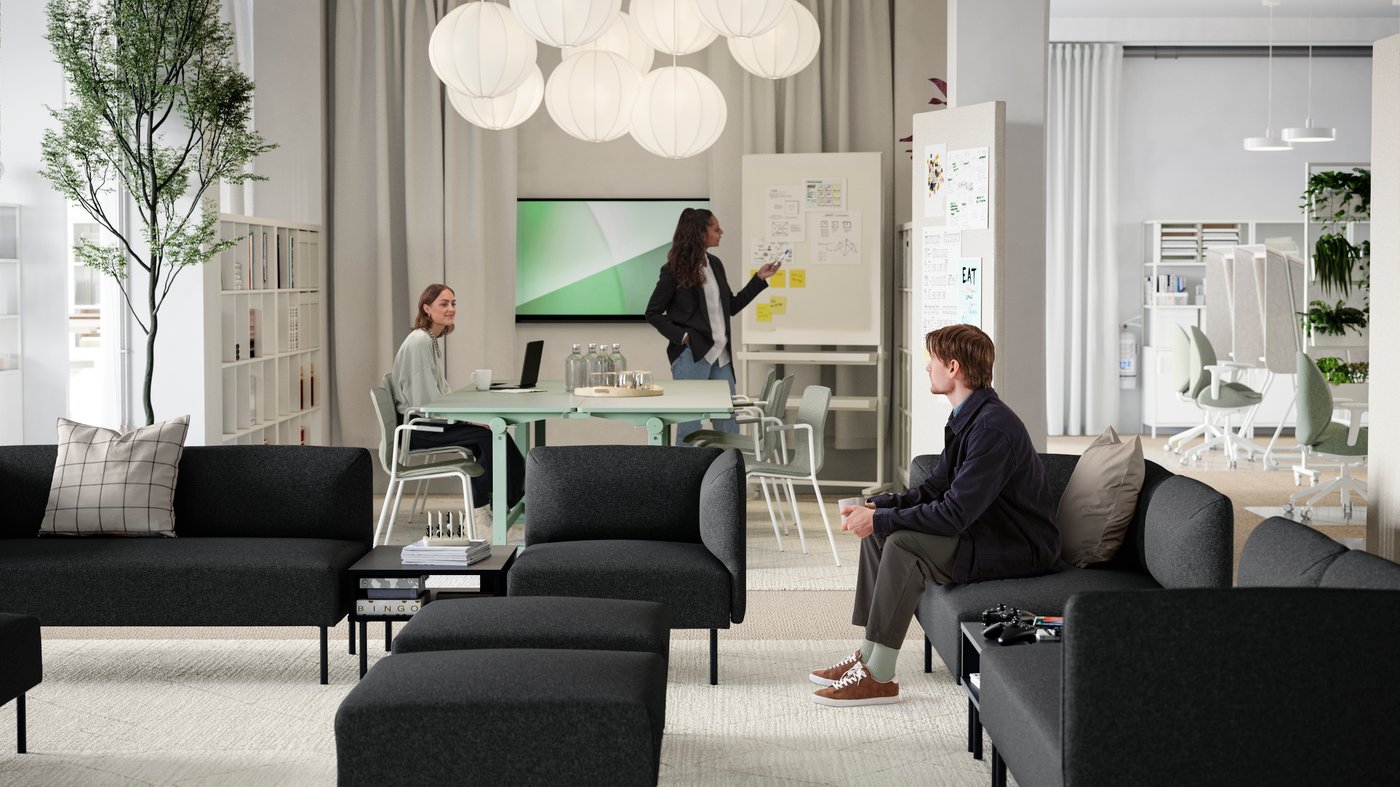 A young man sitting in a lounge area of several dark grey LILLEHEM sofa modules, TUNSTA tables and a meeting area behind it.