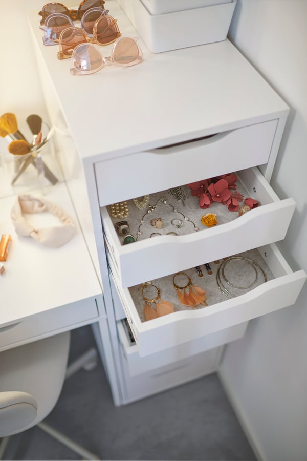 White ALEX drawer unit with two open drawers filled with jewellery such as necklaces, earrings and bracelets. To the left, a white ALEX dressing table with makeup brushes, a hairband and small items.