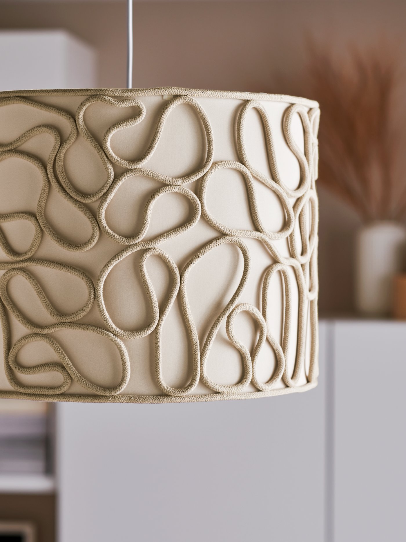 Détails d’une suspension avec abat-jour VINGMAST beige composé de cordons formant des motifs sinueux sur un centre recouvert de textile.