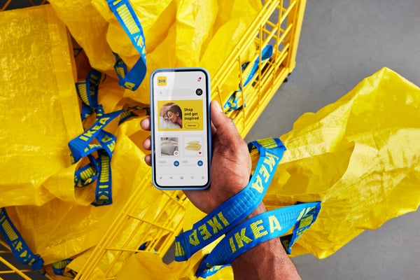 Nečija ruka drži pametni telefon na kom je prikazana IKEA aplikacija, a okolo se nalaze žute IKEA torbe u korpi za kupovinu.