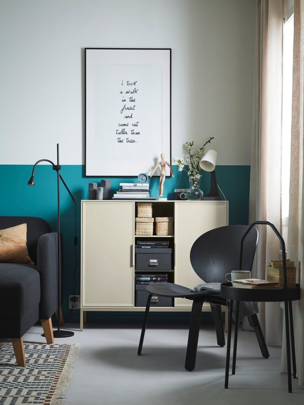 A black FRÖSET easy chair beside a curtained-window with a round black side table beside it holding a cup and a small basket.