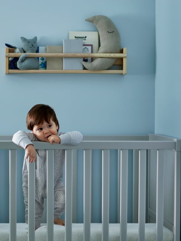 Un lit à barreaux gris SUNDVIK avec un tout-petit qui se tient debout, des livres et des peluches dans un rangement mural à l'arrière.