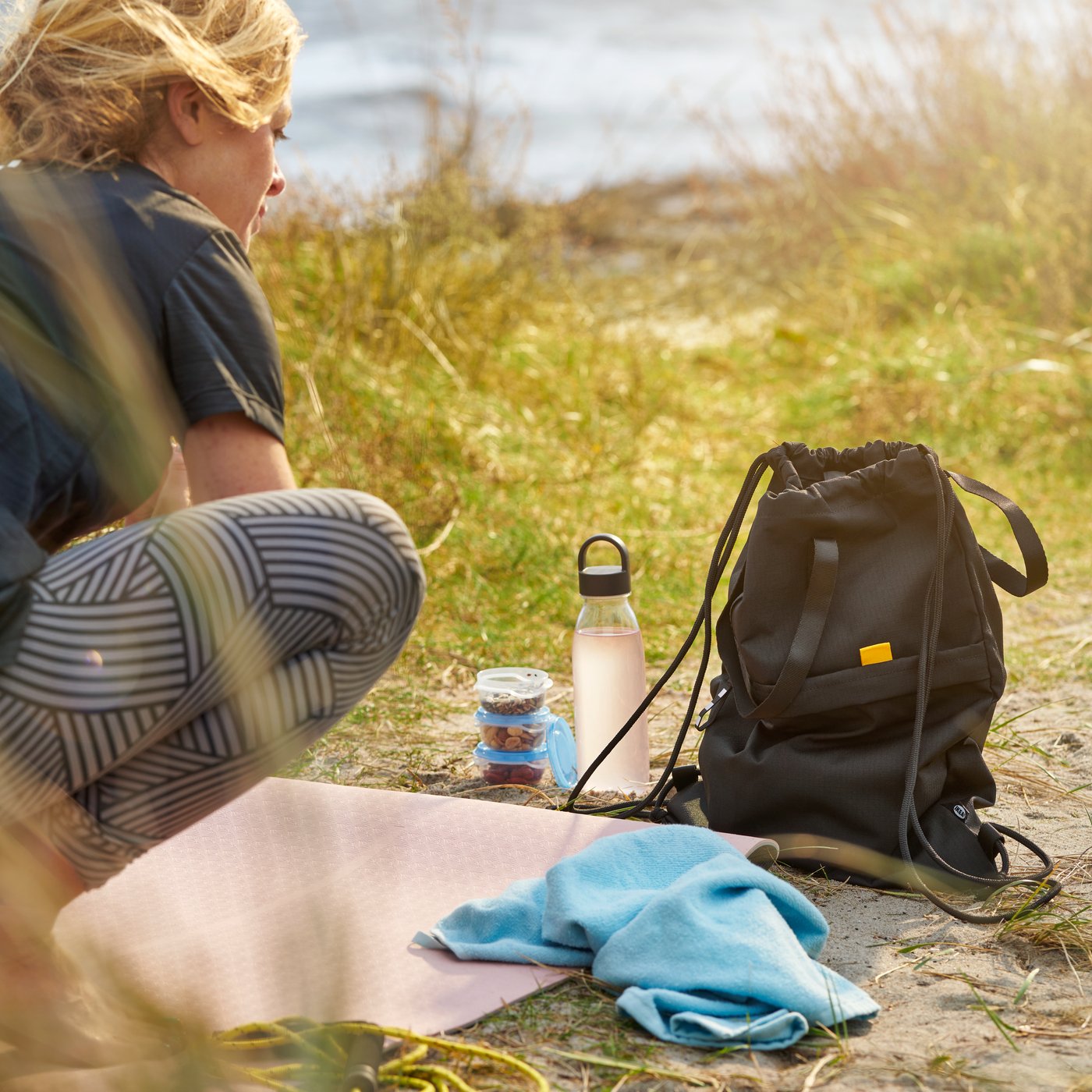 Outdoorová scéna u vody s růžovou podložkou na cvičení, černým sportovním vakem IKEA VÄRMER, modrým ručníkem, sklenicí s oříšky a láhví na vodu IKEA VATTENKRASSE.