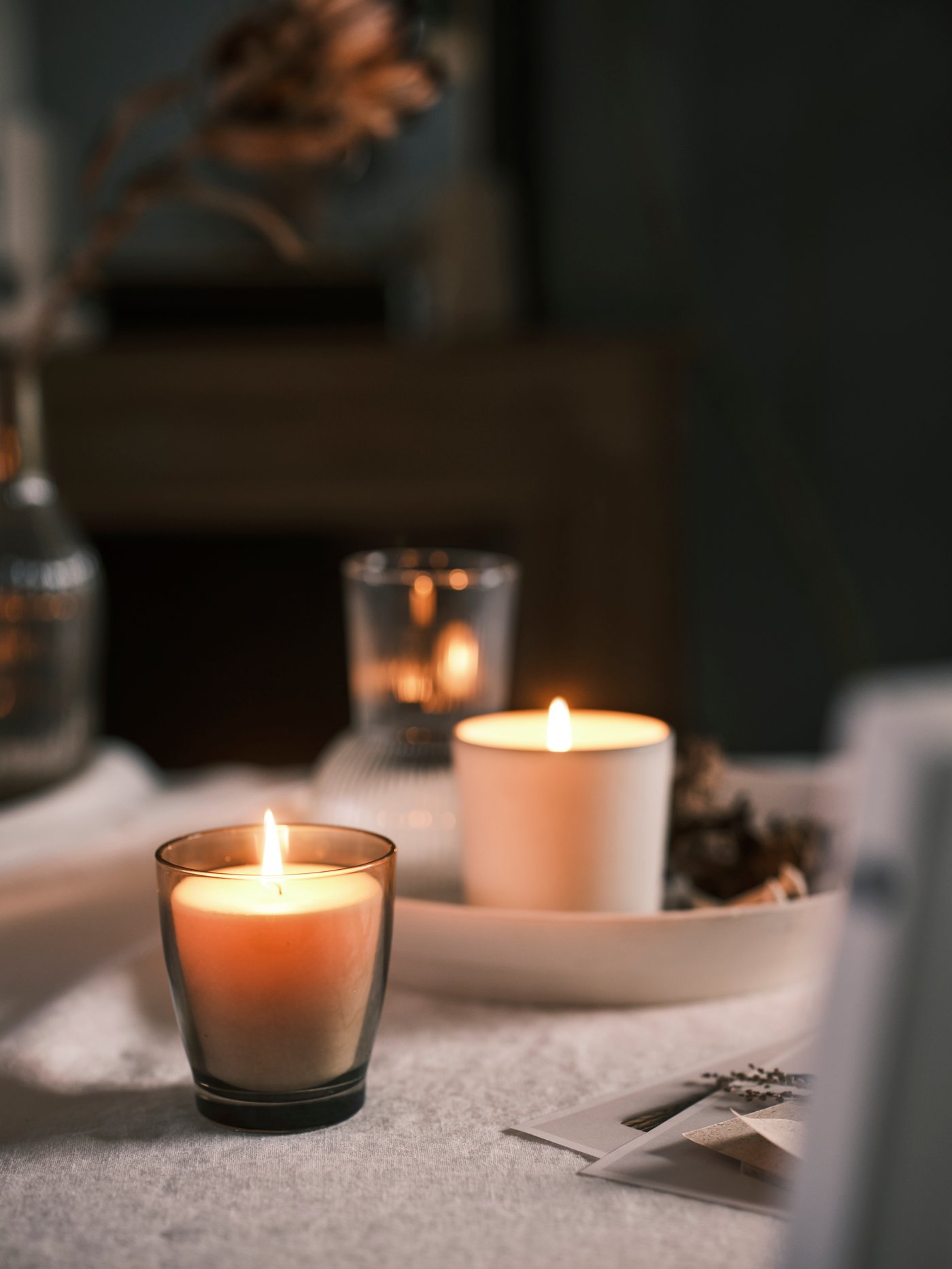 Lilin wangi ENSTAKA berwarna kelabu, lilin wangi ADLAD berwarna putih, hiasan dan piring berwarna putih, semua diletakkan pada alas meja berwarna cerah.