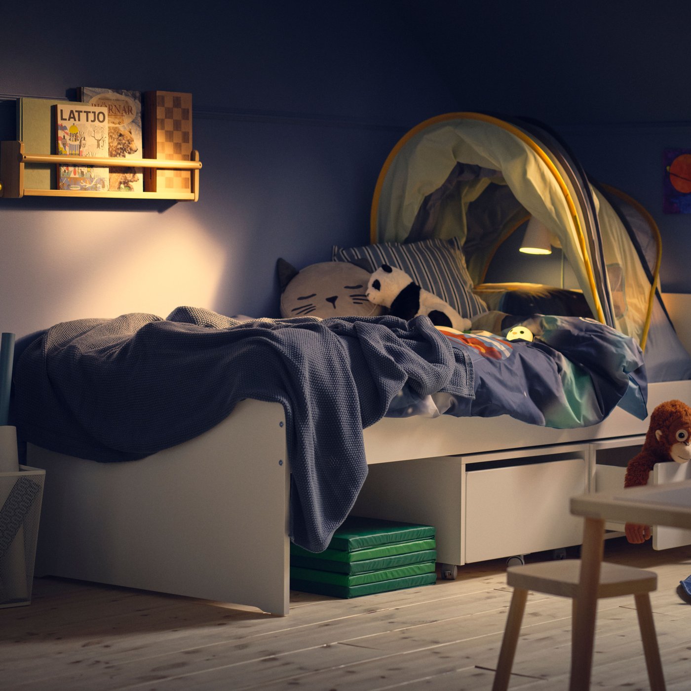 A dim blue children’s room with a white SLÄKT bed with storage boxes with castors underneath and an ELDFLUGA bed tent on top.