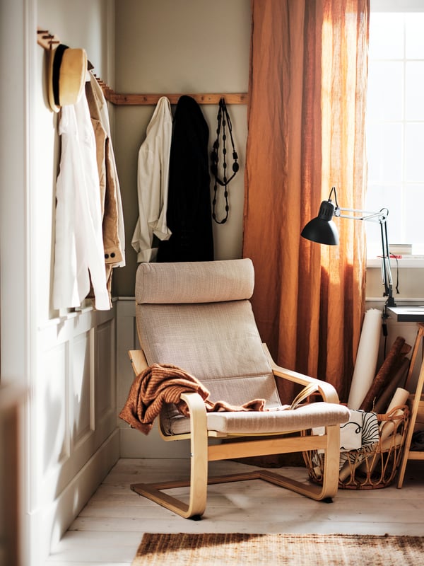 A POÄNG armchair in light beige placed in a corner of a room by a window. An orange curtain and a black desk lamp.