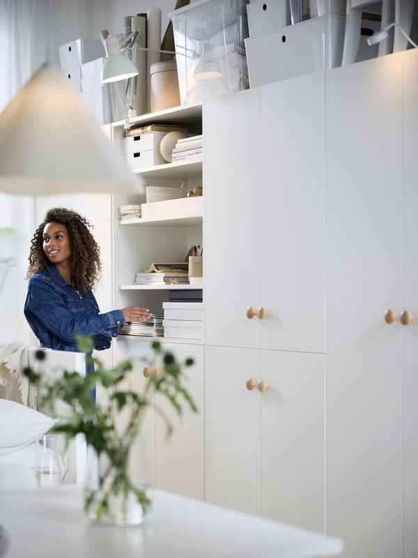 Witte IKEA-opbergwand met het centrale LASTARE-opbergsysteem met open planken en dozen, een vrouw die iets pakt bij de planken, kastdeurtjes met houten knoppen en een onscherpe vaas met groen op de voorgrond.