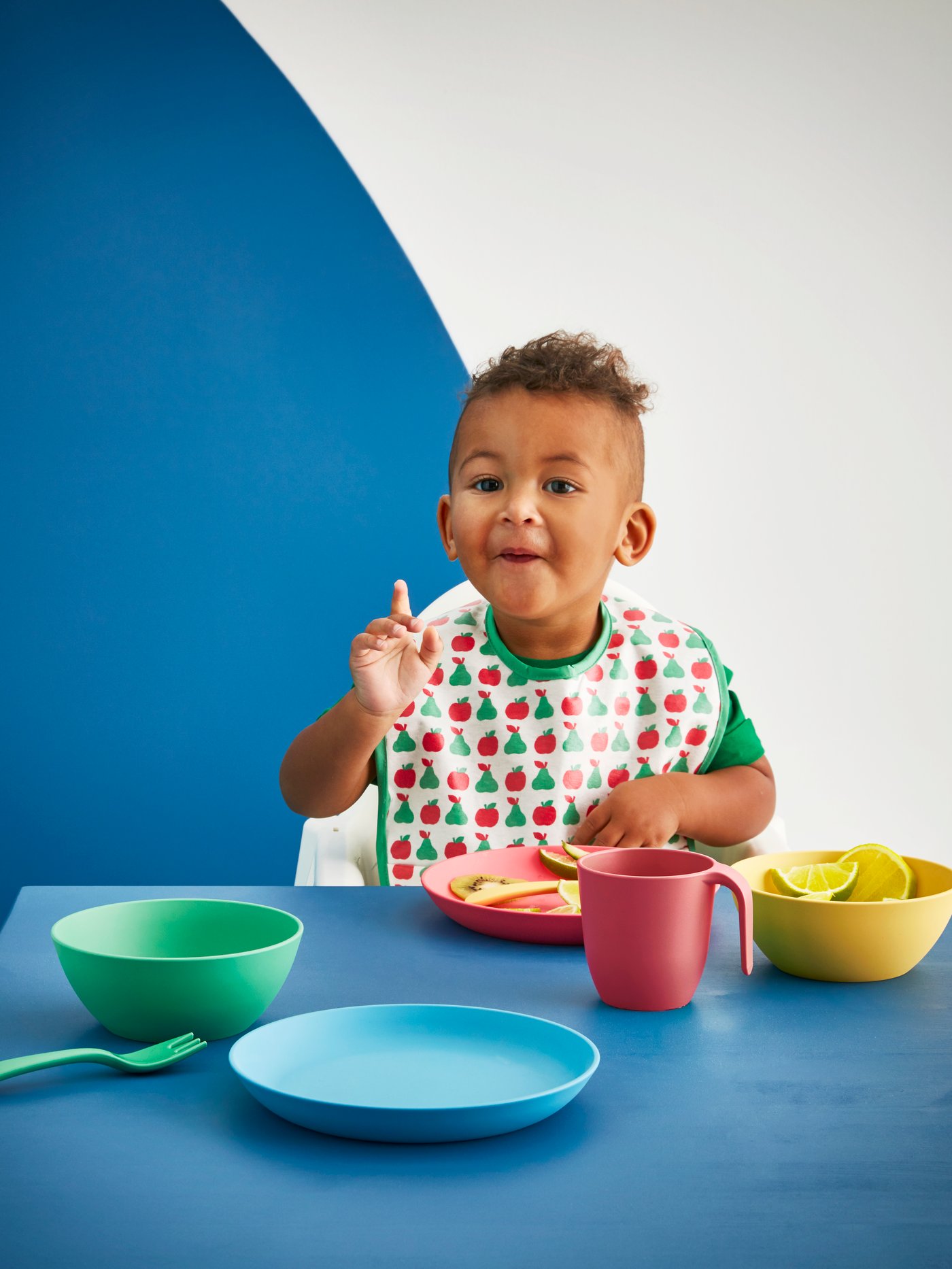 A young boy wearing a MATVRÅ bib sits in a highchair at a table with HEROISK bowls, plates, a mug and cutlery.