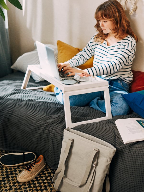 A woman working from bed and typing at a laptop on a white, KLIPSK bed tray, on the floor is a beige, DRÖMSÄCK tote bag.
