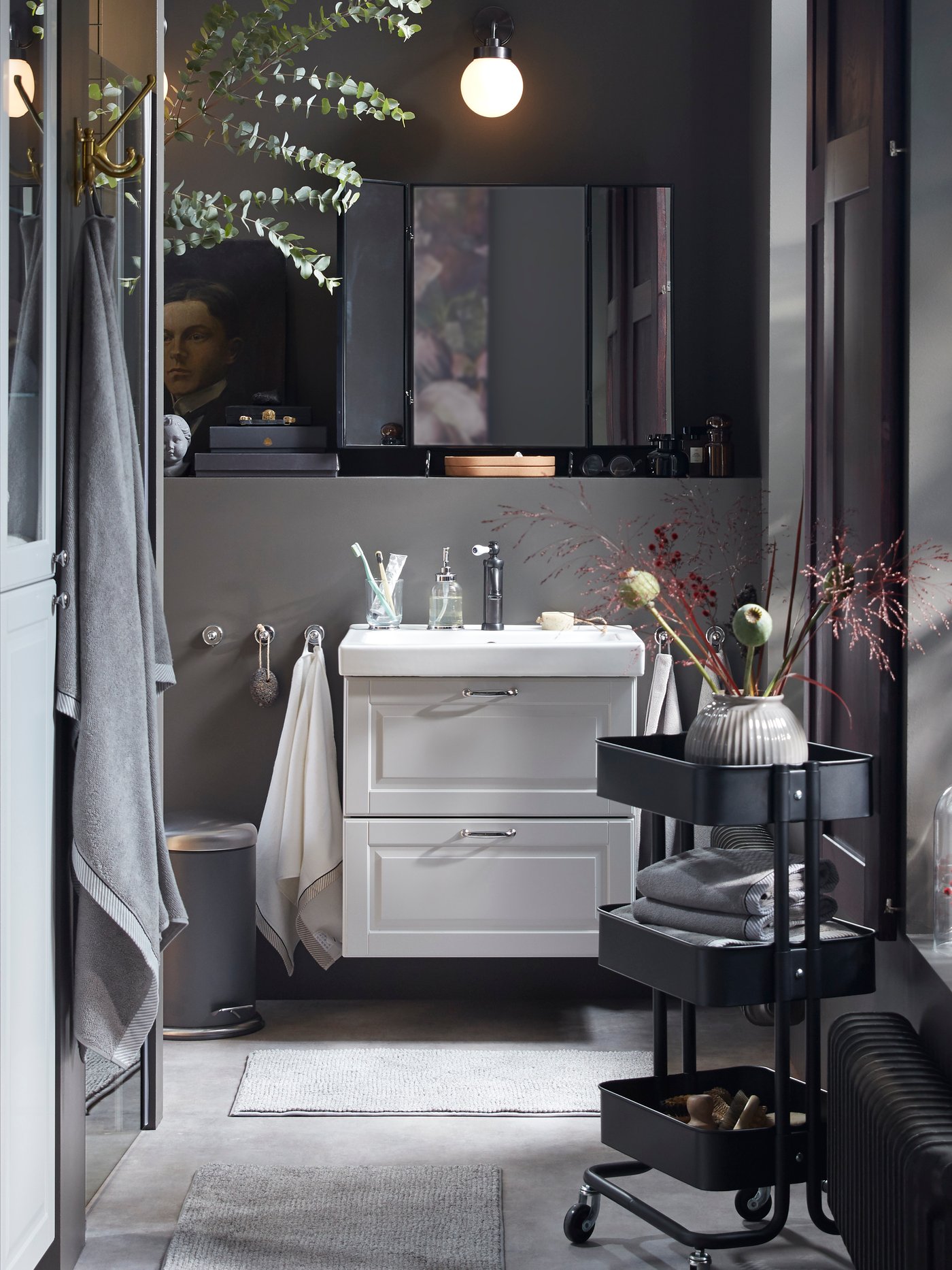 A dark grey bathroom with a wash-stand in light grey, a black RÅSKOG trolley with folded towels and decorative flowers.