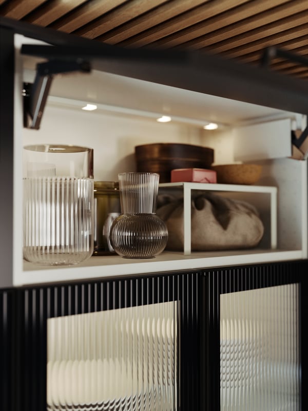 Puertas de cristal HEJSTA en carbón y cristal redondeado e iluminación LED MITTLED en los cajones de la cocina, y un jarrón PÅDRAG en cristal transparente.