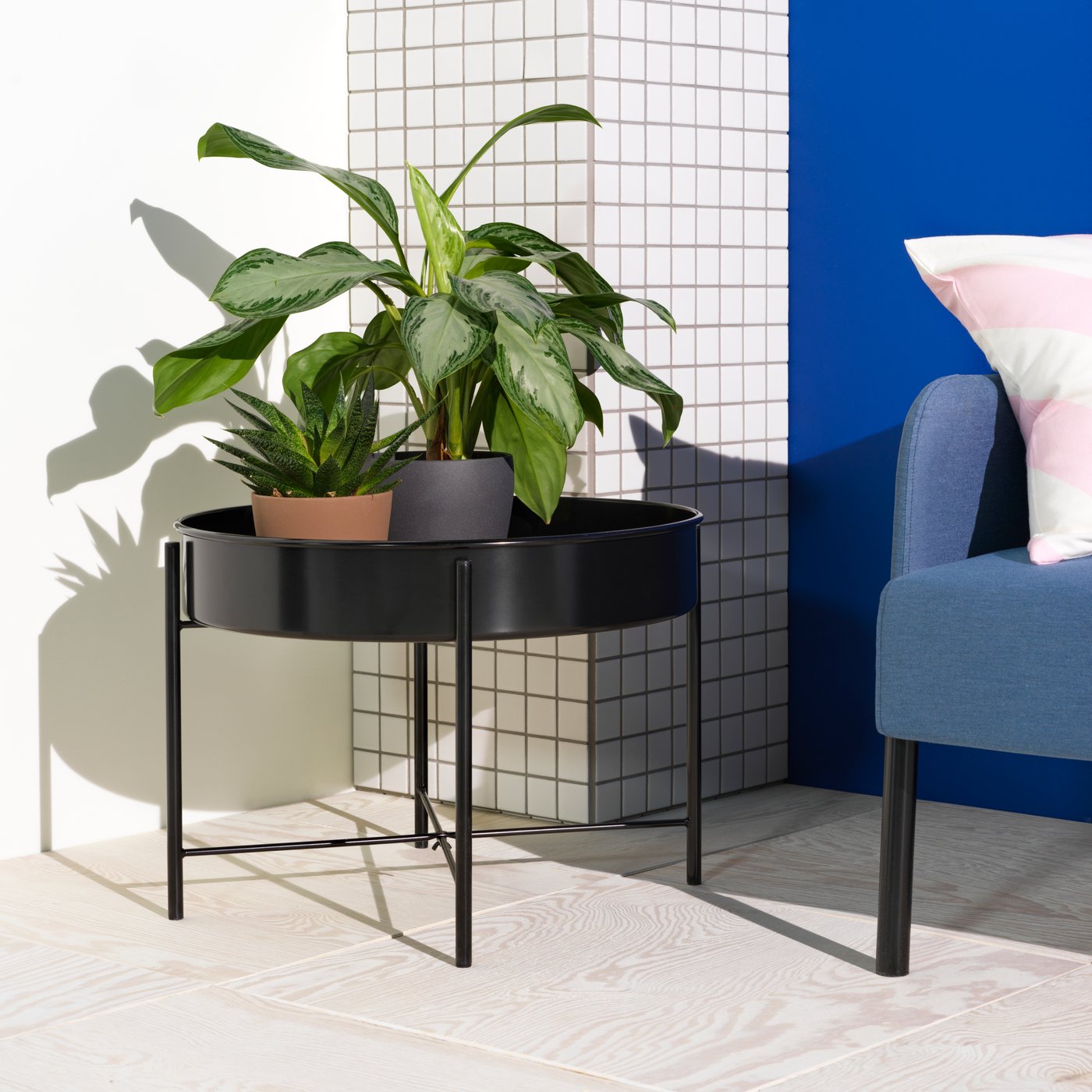 A black KULTURSKOG plant stand with two leafy pot plants is in a bright living room space with a blue sofa.