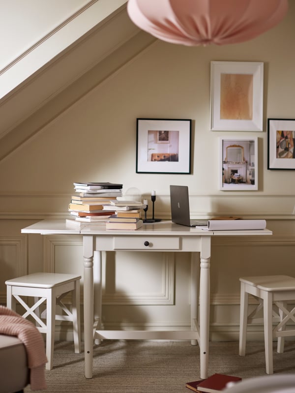 Una mesa blanca plegable INGATORP con libros sobre el tablero, junto a una pared luminosa con cuadros enmarcados y una lámpara rosa de techo encima..