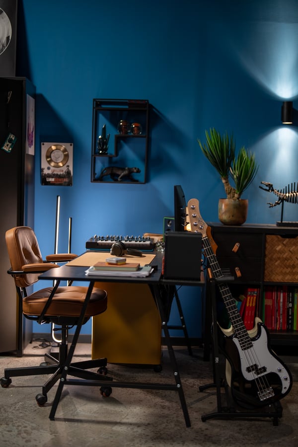 Een compacte werkplek met een IKEA bureau en ALEFJÄLL bureaustoel, aangevuld met wandplanken, een spiegel en muziekaccessoires tegen een blauwe muur.