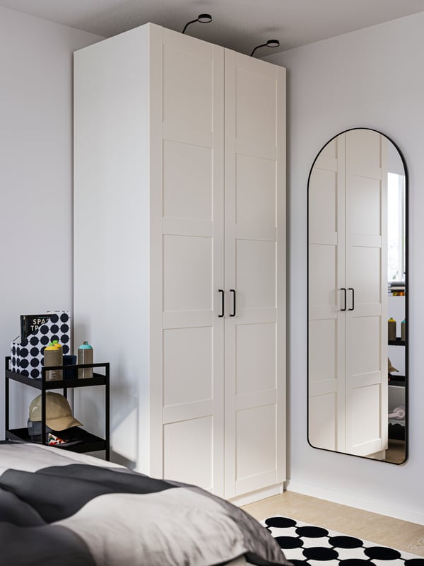 A white PAX wardrobe in the corner of a room, with a LINDBYN mirror on the wall to the right and a bed in the foreground.