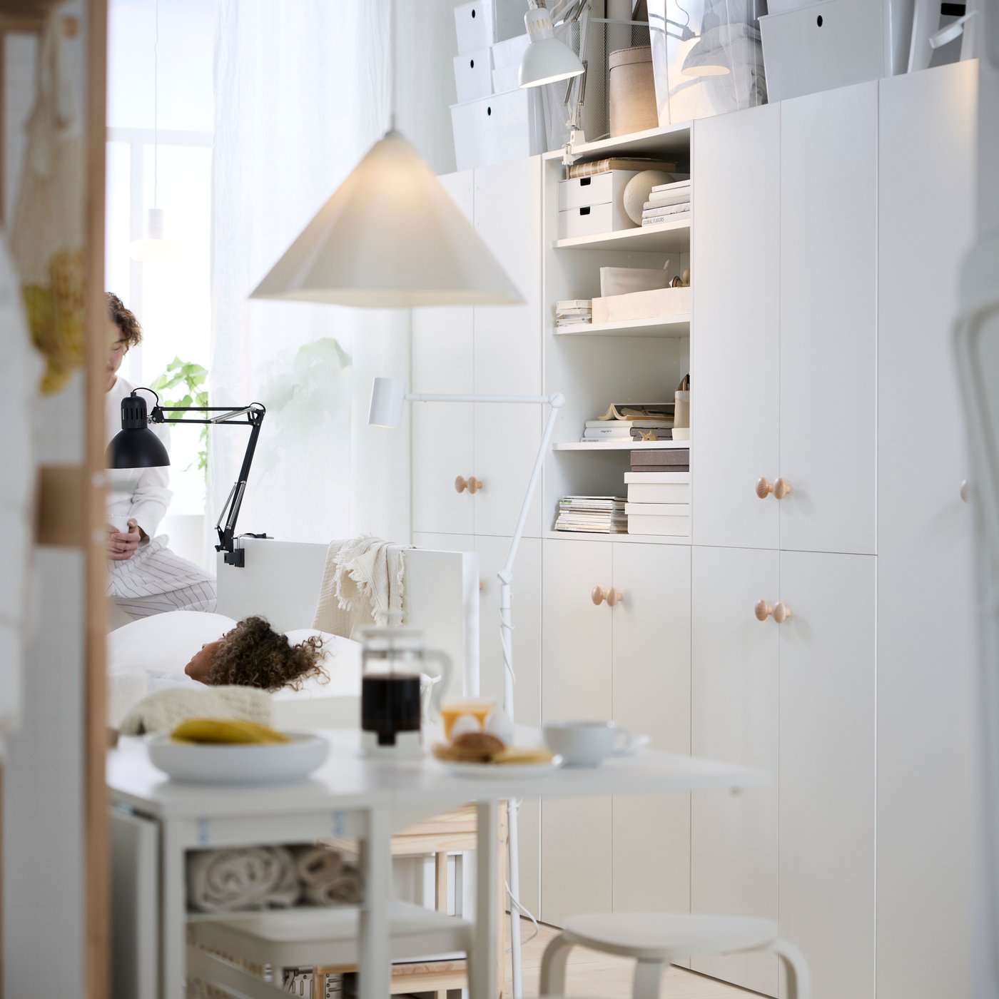 Dans un espace de vie multifonctionnel, rangements LASTARE blancs, cadre de lit blanc et table VIHALS blanche dressée pour le petit déjeuner.