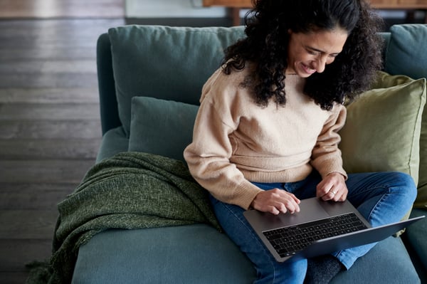 Una donna è seduta su un divano con un computer portatile aperto sulle ginocchia e sorride.