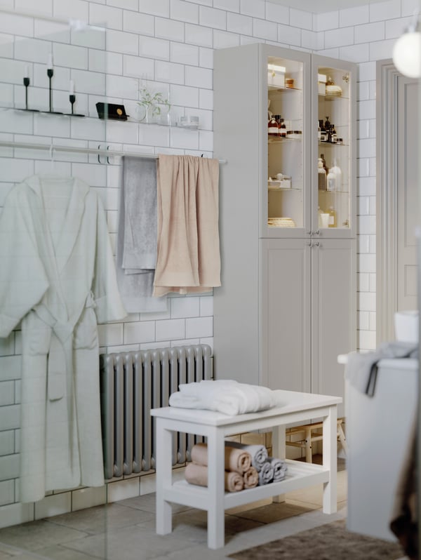 The corner of a tiled bathroom with towels and a bathrobe on two towel rails on the wall beside two GODMORGON high cabinets.