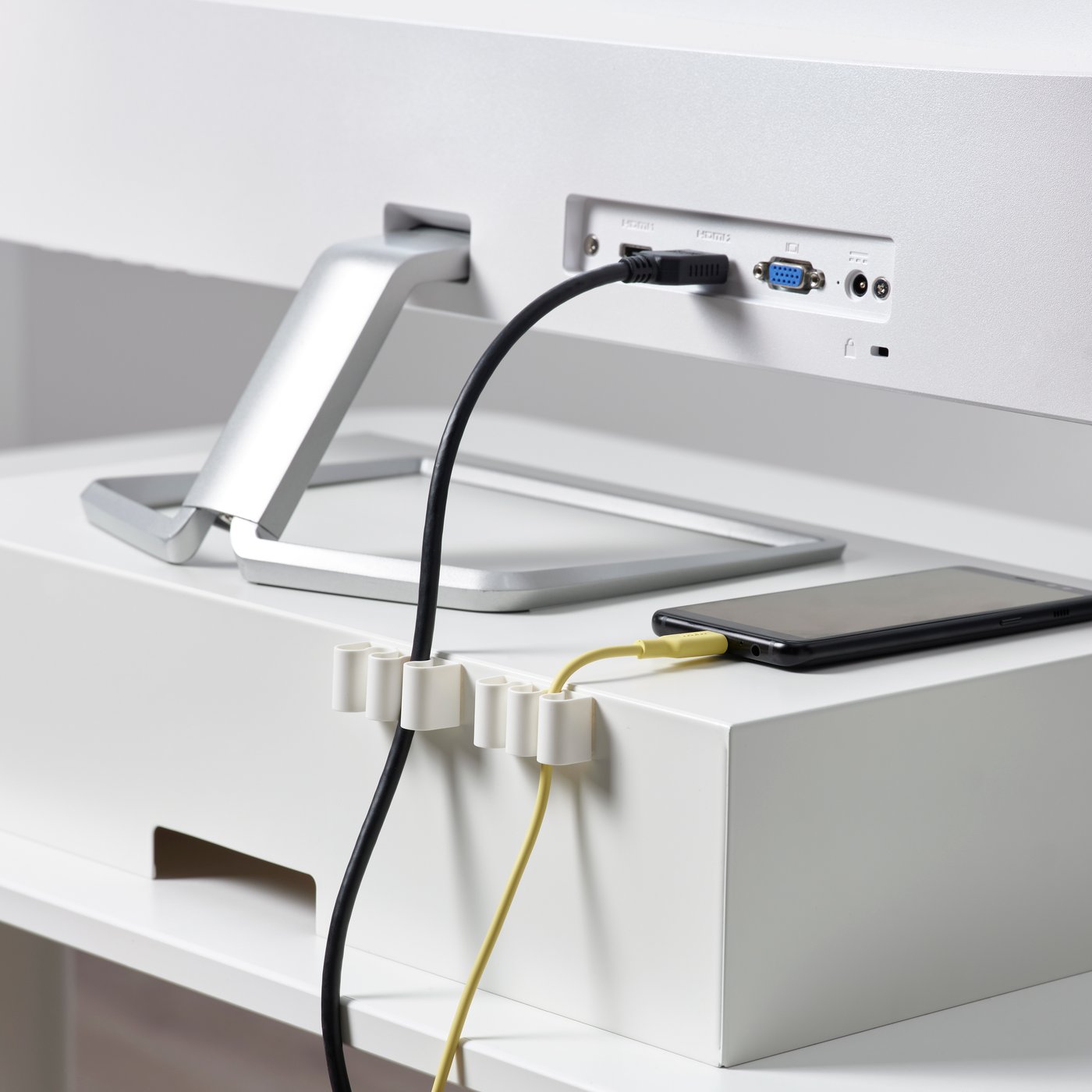 The back of a monitor stand has white plastic KALKSTUFF cable holders on it, keeping a phone cord and an HDMI cable in place.