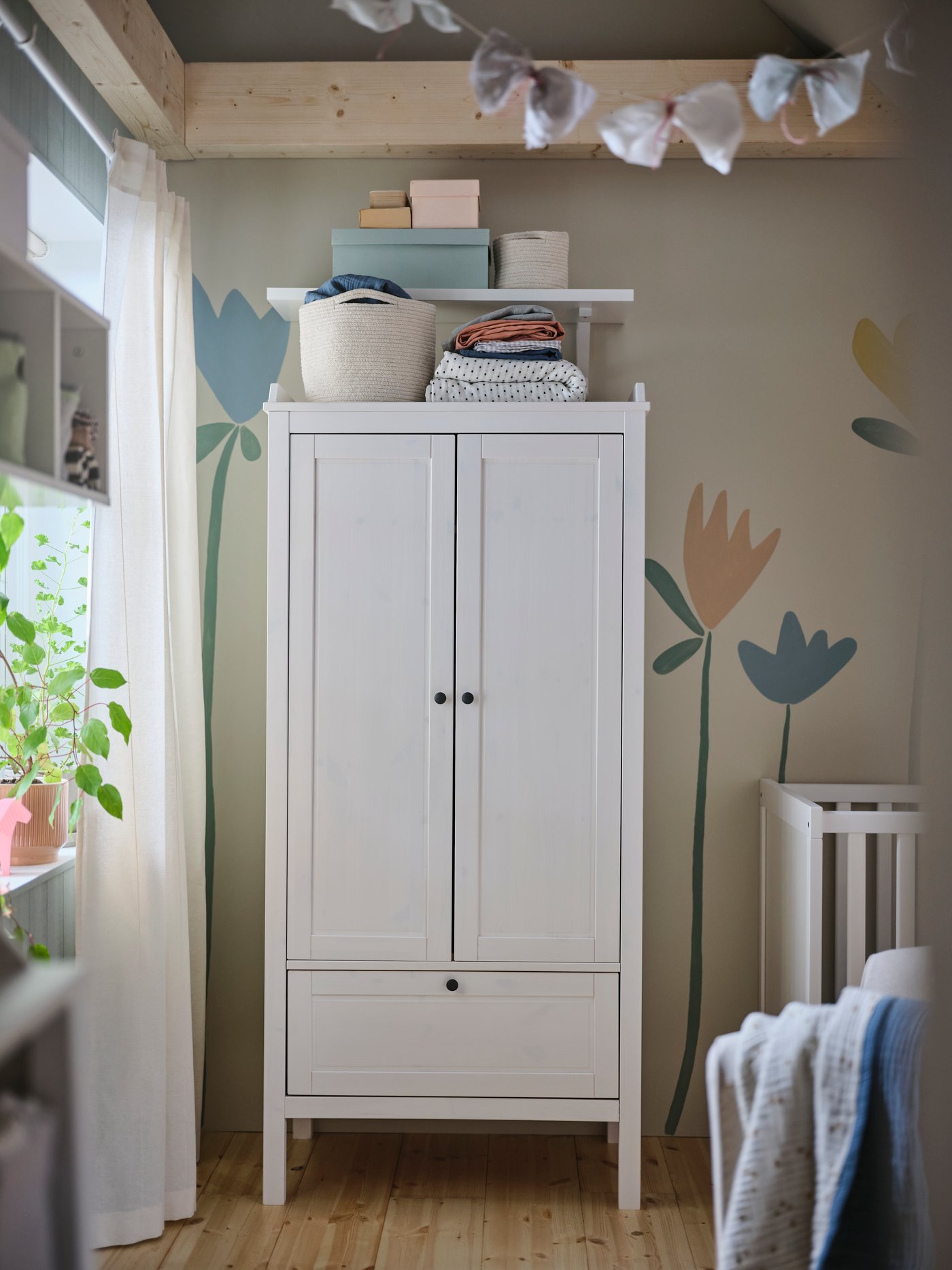 A SUNDVIK wardrobe in a baby’s bedroom with an unbleached cotton TJABBIG basket and a folded blanket on top beside a cot.