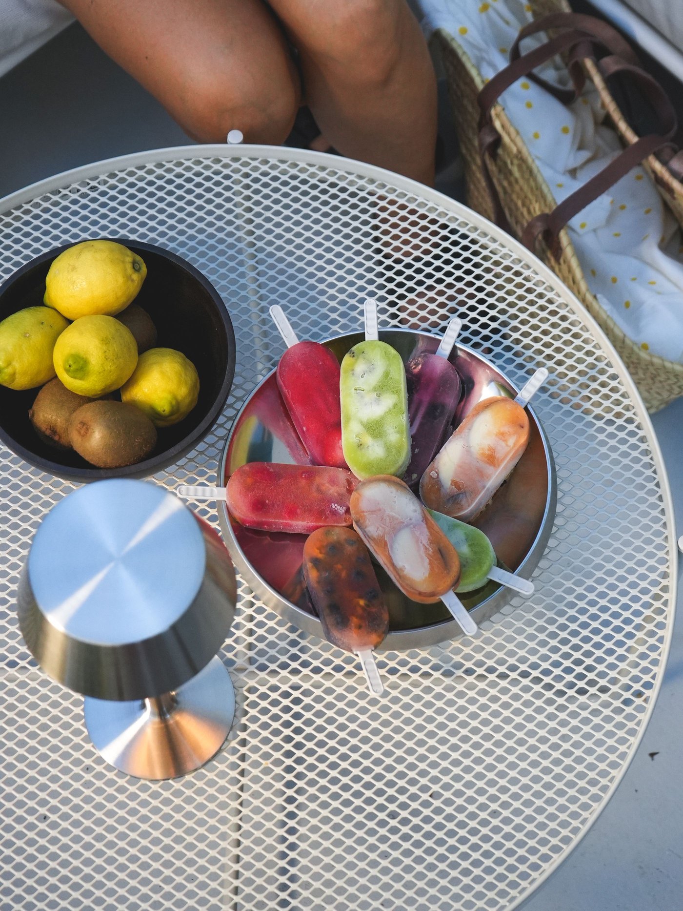 Weisser Metall-Gartentisch mit AVHÅLL Lampe, schwarzer Schale Zitronen/Kiwis, bunte Glace am Stiel.