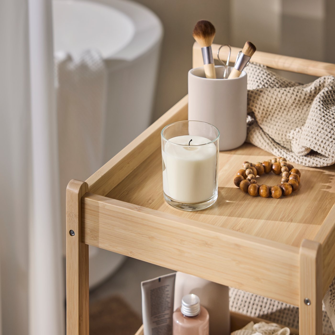 A close-up of a light bamboo STEGESUND trolley displaying a candle, a pot, beige towels, and skincare products in a bathroom.