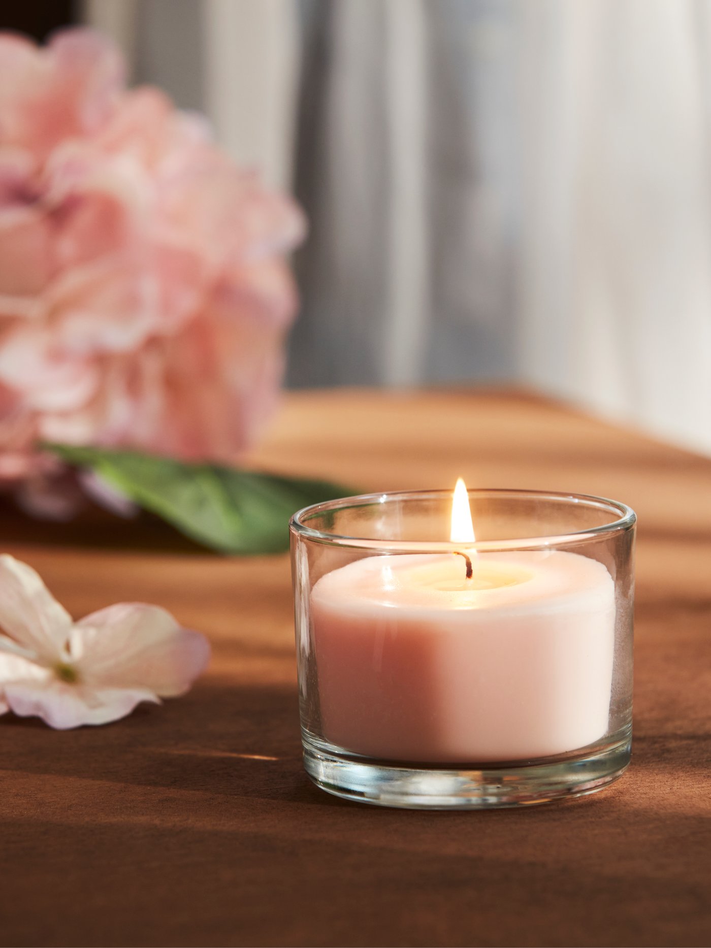 A close-up of a pale pink LOTSFÅGEL scented candle in glass placed on a table with pink flowers in the background.