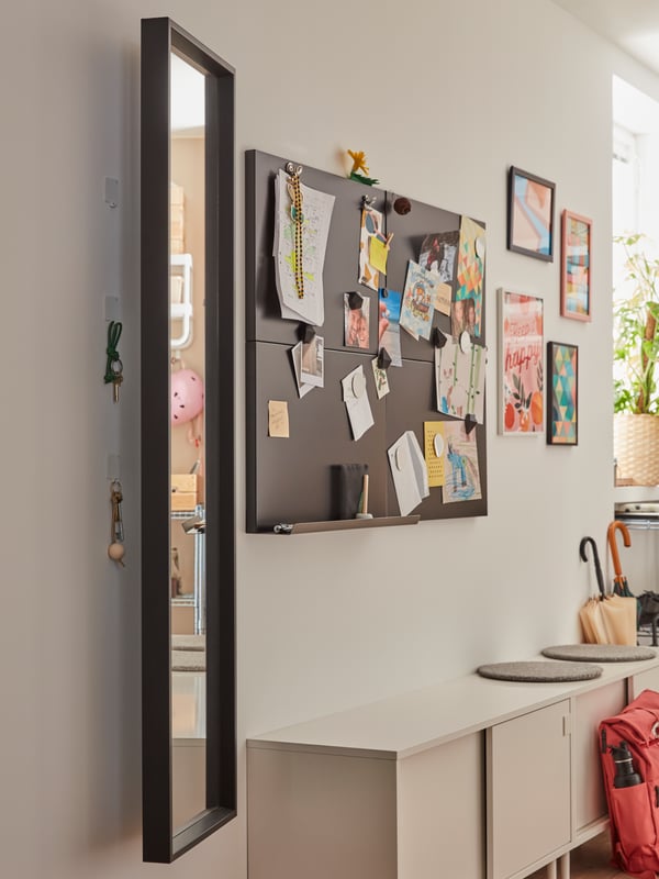 Es ist ein heller Raum mit einem Spiegel und Pin Wänden an der Wand. Daran hängen bunte Bilder und Zettel. Darunter steht eine kleine weiße Kommode.