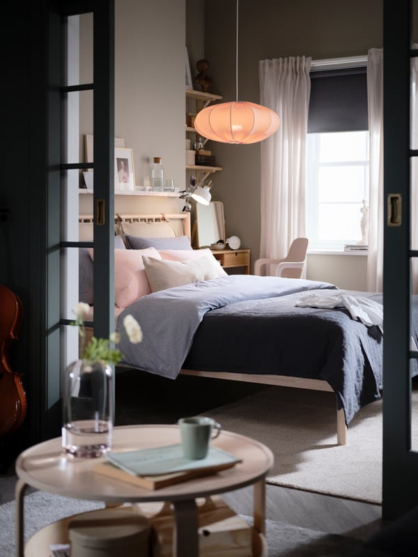 Des portes vitrées entrouvertes s’ouvrent sur une chambre avec un lit BJÖRKSNÄS en bouleau, une coiffeuse et une chaise, et une suspension ovale.