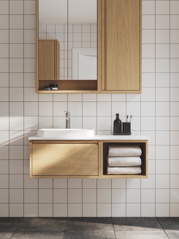 A white-tiled bathroom has an ÄNGSJÖN wash-stand in oak effect with a white semi-recessed wash-basin and a mirror cabinet.